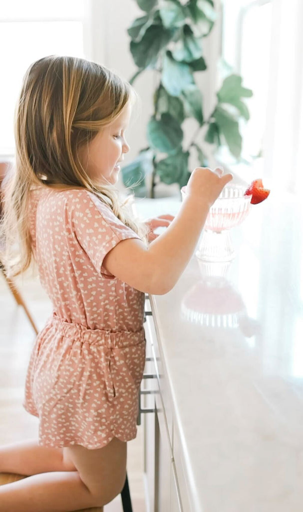 Little girl kneeling on a chair at the kitchen counter, helping prepare a kid-friendly Valentine's mocktail. The girl is wearing a pink and white heart play set from Plain Jane. 